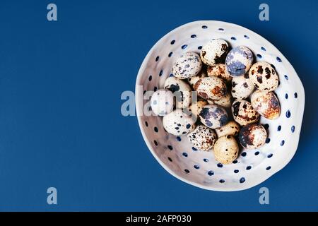 Quail unpainted eggs folded in white ceramic handmade bowl on a blue background Stock Photo
