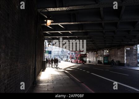 Street scene in Shoreditch, East London with shadows silhouettes of five people walking under a road bridge and red London bus Stock Photo