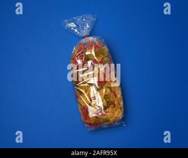 transparent cellophane bag with raw colorful pasta in the form of bows on a blue surface, copy space Stock Photo