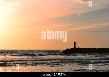 Dramatic sunset by the sea with a red sky reflecting on the sea and the waves behind the silhouette of a lighthouse on the coast Stock Photo