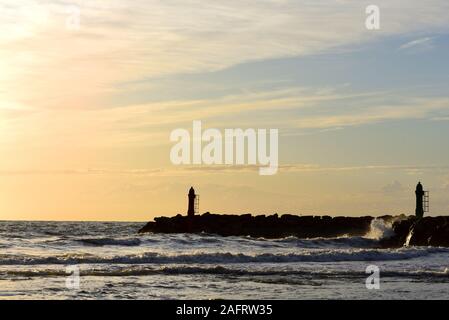 Dramatic sunset by the sea with a red sky reflecting on the sea and the waves behind the silhouette of two small lighthouses on the coast Stock Photo