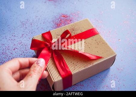 Woman's hand with beautiful manicure holding a giftbox tied with bright red color ribbon on purple background with sparklets and confetti. Stock Photo