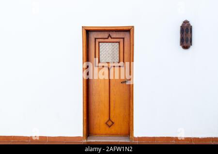 Old wooden door in arabic style. White wall with a door in the middle. A lantern on the side on a white wall near the door. Conceptual minimalism.