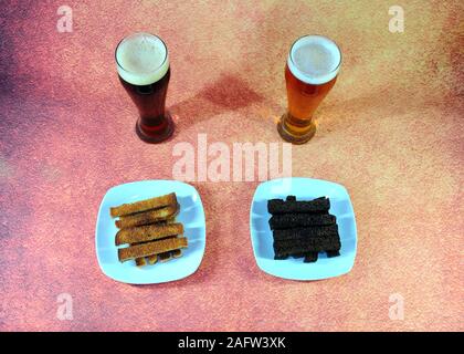 Two tall glasses with light and dark beer and two white ceramic plates with wheat and rye croutons, on a brown abstract background. Close-up. Stock Photo