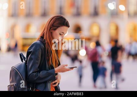 Young woman with backpack using smart phone on urban sidewalk Stock Photo