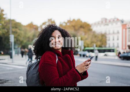 Smiling young woman with smart phone looking over shoulder on city street Stock Photo