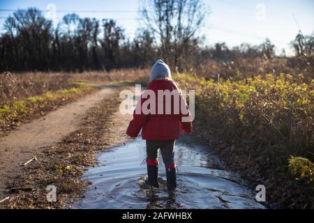 3 years old child splashing in a puddle with red jacket and wellies in a country road on a sunny winter day. Back view.
