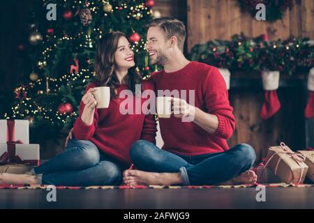 Photo of cute couple spending newyear night in decorated garland lights room sitting cozy on floor near evergreen tree drinking hot beverage indoors Stock Photo