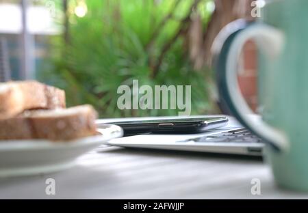 Working on the computer and cell phone while having breakfast. Work from home or flexi time concept. Stock Photo