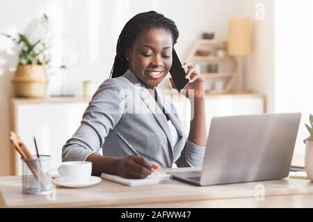 Afro Businesswoman Taking Notes While Having Phone Conversation In Modern Office Stock Photo