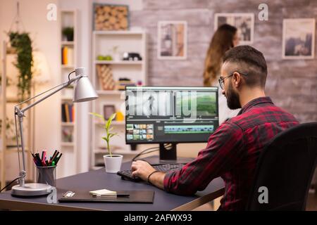 Back view of creative filmmaker working on a movie on laptop. Girlfriend in the background. Stock Photo