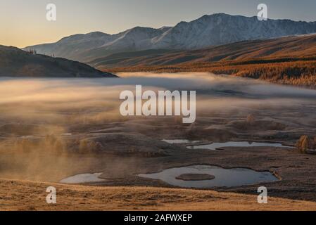 Amazing autumn view with mountains covered with snow and a golden forest, white fluffy fog over the lakes, valley and grass in hoarfrost at sunrise