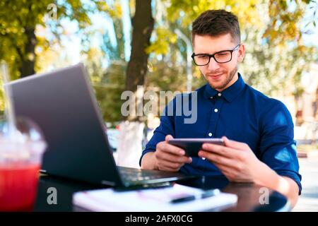 Handsome businessman distracted from work on the laptop watching video on smartphone. Freelancer holding mobile phone and browsing using high speed 4g Stock Photo