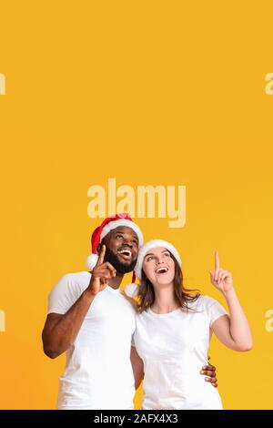 Excited interracial couple in Santa hats pointing upwards at copy space Stock Photo