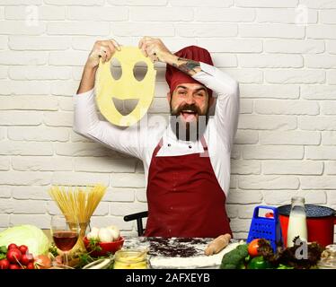 Man with beard holds dough with cut smiley on white brick background. Cook with excited face in uniform sits by table with vegetables and kitchenware. Professional cookery concept. Chef makes dough Stock Photo