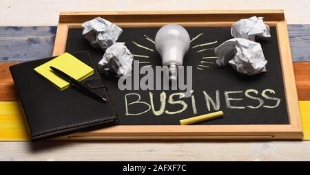 Process of creation: lamp, crumpled paper, chalk and notebook. Board with light bulb, yellow chalk, pen and stickers. Chalkboard with business accessories on wooden background. Business idea concept Stock Photo