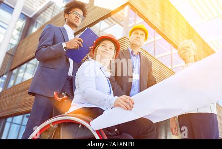 Disabled woman in a wheelchair as an architect with blueprint next to engineers and business people Stock Photo