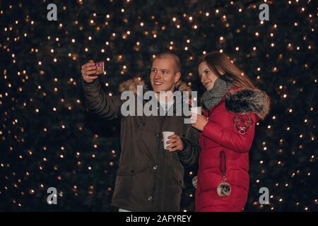 Happy young couple making selfie and smiling while holding cup of mulled wine and standing in front of a Christmas background Stock Photo