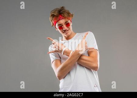 stylish blond guy with a bandana in glasses and a white T-shirt crossed his arms and shows a rocker sign on a gray background Stock Photo