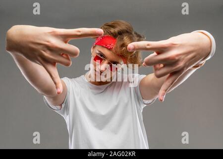 European blond guy with a bandana in glasses and a white T-shirt shows a rocker sign with his hands forward on a gray background Stock Photo