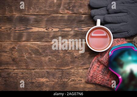 Snowboard googles, cup of tea and sport gloves on brown wooden table background with copy space. Stock Photo
