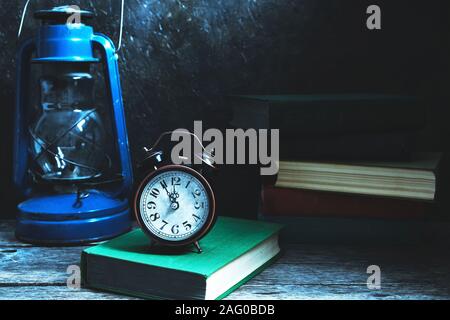 An old vintage alarm clock on a table among books and a kerosene lamp in the moonlight. Stock Photo