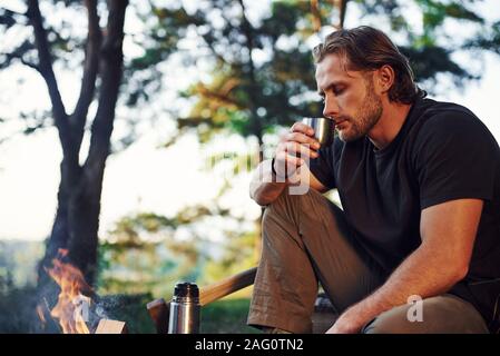 Making drink by using thermos. Man in black shirt near the campfire in the forest at his weekend time Stock Photo