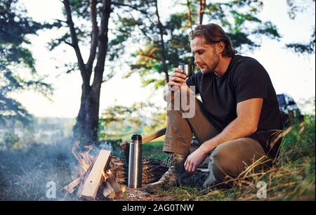 Making drink by using thermos. Man in black shirt near the campfire in the forest at his weekend time Stock Photo