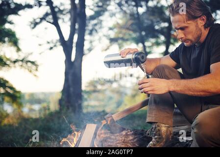 Making drink by using thermos. Man in black shirt near the campfire in the forest at his weekend time Stock Photo