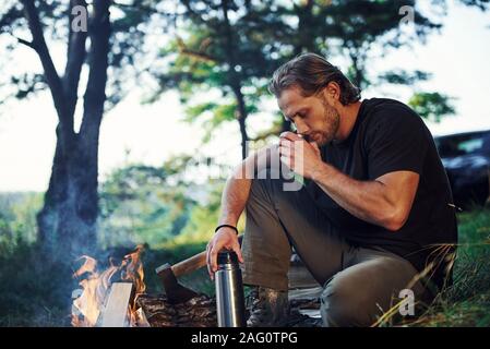 Making drink by using thermos. Man in black shirt near the campfire in the forest at his weekend time Stock Photo