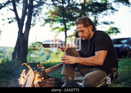 Making drink by using thermos. Man in black shirt near the campfire in the forest at his weekend time Stock Photo