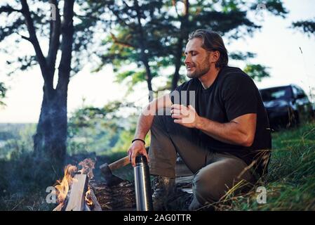 Making drink by using thermos. Man in black shirt near the campfire in the forest at his weekend time Stock Photo