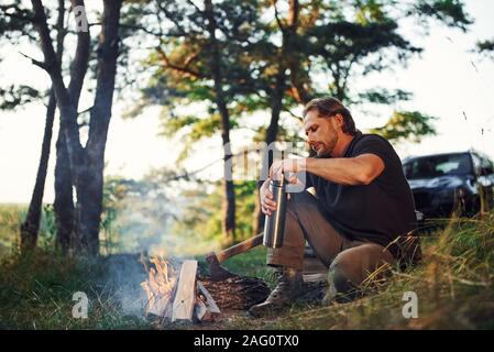 Making drink by using thermos. Man in black shirt near the campfire in the forest at his weekend time Stock Photo