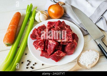 Raw ingredients for cooking stewed meat or goulash: pieces of fresh beef, carrot, onions, garlic, celery and salt on white wooden table. Cook at home. Stock Photo
