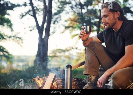 Making drink by using thermos. Man in black shirt near the campfire in the forest at his weekend time Stock Photo