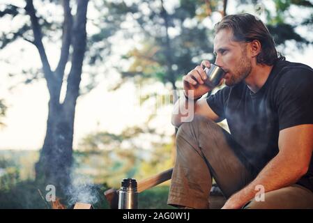 Making drink by using thermos. Man in black shirt near the campfire in the forest at his weekend time Stock Photo