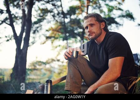 Making drink by using thermos. Man in black shirt near the campfire in the forest at his weekend time Stock Photo