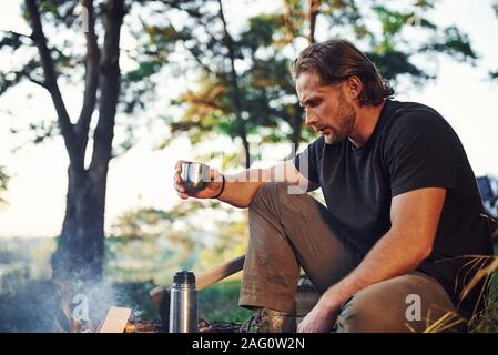 Making drink by using thermos. Man in black shirt near the campfire in the forest at his weekend time Stock Photo