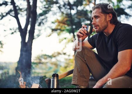 Making drink by using thermos. Man in black shirt near the campfire in the forest at his weekend time Stock Photo