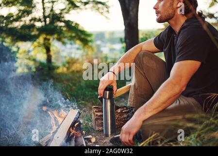 Making drink by using thermos. Man in black shirt near the campfire in the forest at his weekend time Stock Photo