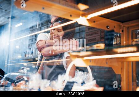 Embracing each other. Behind the glass. Light stripes. Two young cafe workers indoors. Conception of business and service Stock Photo