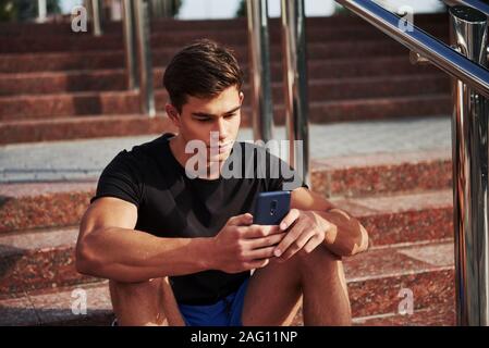 Young guy in casual clothes sits on the stairs with smartphone in hands Stock Photo