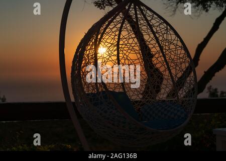 Hanging cane swing chair providing theme of relaxation in summer silhouetted by setting sun Stock Photo