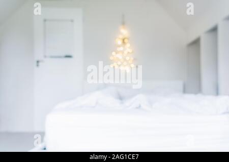 Blurred background of glowing lights Christmas tree on the white wall above the bed in the bedroom. Simple, minimal conscientious Christmass interior Stock Photo