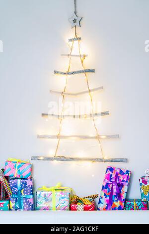 Vertical background of Unusual Christmas tree made of wooden sticks with festive lights on the white wall and colourful gift boxes. Simple, minimal co Stock Photo