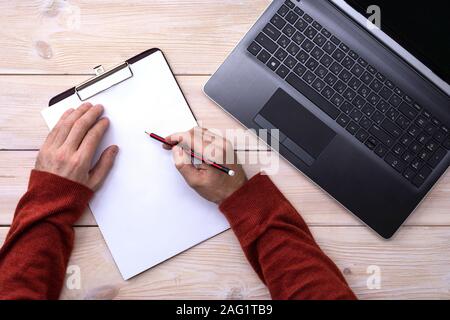 A man works on a laptop, hands write in a notebook with a pencil on a wooden table, top view Stock Photo