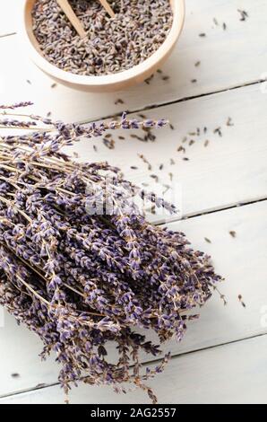 Bunch of dried lavender flowers in foreground with bowl of loose buds in background. Also scattered on white wood plank table.. Stock Photo