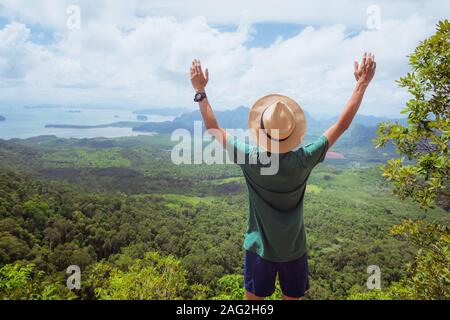 Stock image: a happy traveler with his hands raised up in admiration stands on top of high mountain and watches landscape of forest, hills, and sea.