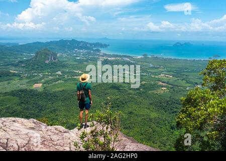 Traveler in hat stands on rock high in mountains landscape & sea on horizon. View of Ao Nang bay and islands from Dragon Crest at Khao Ngon Nak Trail Stock Photo