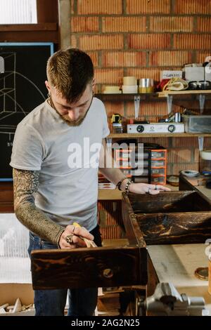 Tattooed artist applying varnish paint on a wooden furniture at craft workshop Stock Photo